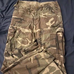 Cargobyxor i camouflage från Japan Rags - Säljer ett par cargobyxor i camouflage från Japan Rags. Byxorna har flera stora fickor på sidorna, knäppning med knappar och klassisk cargomodell. Perfekta för en avslappnad och cool stil.