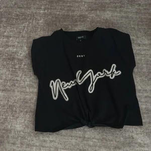 Svart t-shirt från DKNY med New York-tryck - Svart t-shirt från DKNY med vit text 'New York' framtill. Klassisk rund halsringning och korta ärmar. Perfekt för en avslappnad och trendig look.