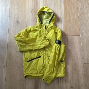 Gul vindjacka från Stone Island - Snygg gul vindjacka från Stone Island med huva och svarta detaljer. Jackan har dragkedja framtill, två snedställda fickor med dragkedja och klassisk logotyp på ärmen. 