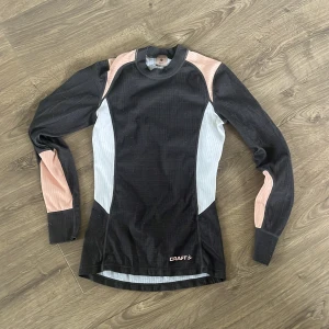 Svart långärmad träningströja från Craft - Säljer en svart långärmad träningströja från Craft med ljusgrå och ljusrosa paneler på sidorna och ärmarna. Perfekt för löpning eller annan träning. Tight passform och hög hals.