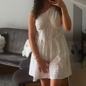 Studentklänning - Säljer en söt vit, kort klänning med one shoulder-design och broderade detaljer. Den är från Asos och går ej att köpa på hemsidan längre. Endast använd en gång och i mycket bra skick. Klänningen är dessutom inte genomskinlig vid bysten. Perfekt till studenten!🤍