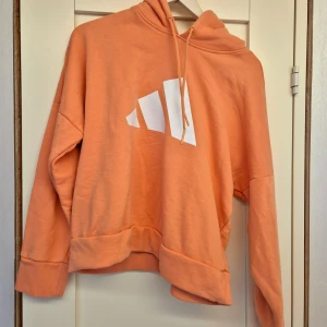 Persikofärgad hoodie från Adidas - Säljer en persikofärgad hoodie från Adidas med vit logga framtill. Tröjan har en oversized passform, huva med dragsko och långa ärmar. Perfekt för en avslappnad stil och riktigt skön att bära. Storlek M barn 12-14år