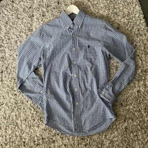Blårutig skjorta från Ralph Lauren - Säljer en klassisk blårutig skjorta från Ralph Lauren i slim fit-modell. Skjortan har långa ärmar, knappar framtill och broderad logga på bröstet. Perfekt för dig som gillar stilrena och tidlösa plagg.