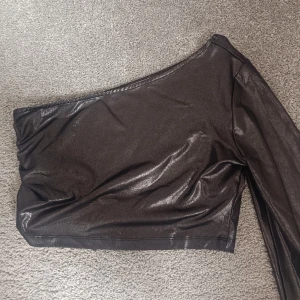 Oneshoulder topp från NA-KD - Säljer en svart/ grå/ silvrig glansig oneshoulder topp från NA-KD i storlek S. Toppen har en lång vid ärm och är tillverkad i ett stretchigt syntetmaterial som ger en snygg och modern look. Väldigt snygg och passar perfekt som fest topp. 90 kr men priset kan diskuteras!! 