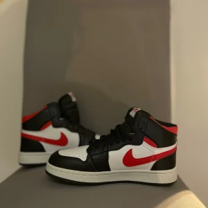 Nike Air Jordan 1 Mid med röd swoosh - Säljer ett par Nike Air Jordan 1 Mid med svart, vitt och rött färgschema. Skorna har klassisk hög siluett, svart snörning och röd swoosh på sidorna. Perfekta för dig som gillar streetwear och vill sticka ut med en ikonisk sneaker.
