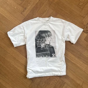 Vit t-shirt från Maison Margiela - Säljer en vit t-shirt från Maison Margiela med ett stort tryck på framsidan. Trycket har en svartvit bild och text med siffror och varumärkesnamnet. Perfekt för dig som gillar stilrena och unika plagg. Självklart äkta