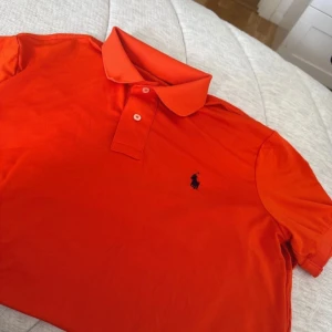 Orange pikétröja från Ralph Lauren - Säljer en orange pikétröja från Ralph Lauren med klassisk krage och två knappar framtill. Tröjan har korta ärmar och den ikoniska broderade loggan på bröstet. Perfekt för en sportig och stilren look.