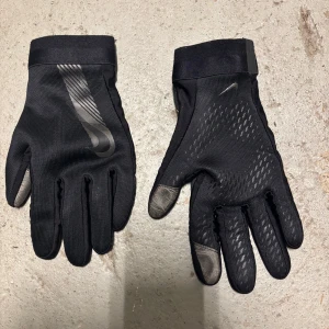 Svarta träningshandskar från Nike - Svarta handskar från Nike med diskret mönster och logga på ovansidan. Handskarna har förstärkta fingertoppar och greppvänligt material i handflatan. Perfekta för träning utomhus under kyligare dagar.