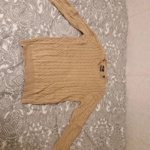 Beige kabelstickad tröja från Gant - Säljer en beige kabelstickad tröja från Gant i premium bomull. Tröjan har rund halsringning och långa ärmar, perfekt för lager-på-lager. Klassisk och stilren design med diskret logga på bröstet. Använd fåtal gånger