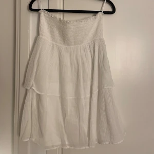 Vit axelbandslös klänning med volang - Säljer en söt vit axelbandslös klänning med smock upptill och volangig kjol. Klänningen är luftig och har en lätt, flowy känsla – perfekt för varma dagar eller sommarens fester.
