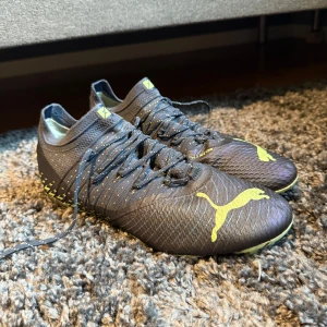 Mörkblåa fotbollsskor från Puma med gröna detaljer - Säljer ett par blåa fotbollsskor från Puma med gröna detaljer och logga. Skorna har snörning och mönstrad sula med dobbar i grön och blå blixtmönster. Perfekta för dig som vill sticka ut på planen!
