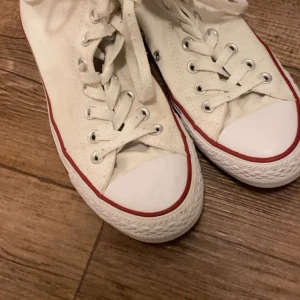 Vita Converse All Star high-top sneakers - Klassiska vita Converse All Star high-top sneakers med röd och blå rand längs sulan. Skorna har snörning och rund tå, samt Converse-loggan på sidan. Perfekta för en avslappnad och tidlös stil.