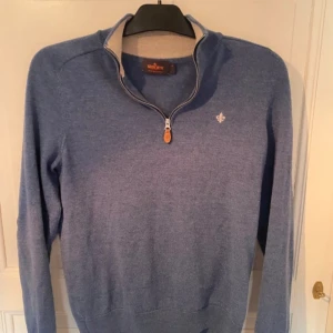 Blå merinoulltröja med dragkedja från Morris - Säljer en blå långärmad tröja från Morris i 100% merinoull. Tröjan har halv dragkedja framtill och en liten broderad logga på bröstet. Perfekt för lager-på-lager och snygg till jeans eller chinos.