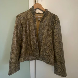 Ormmönstrad jacka från Kikit Studio - Säljer en snygg jacka från Kikit Studio med ormmönster i beige och bruna toner. Jackan har dragkedja framtill, hög krage och lång ärm. Materialet är syntet med nylonfoder. Perfekt för dig som vill sticka ut med en cool look.