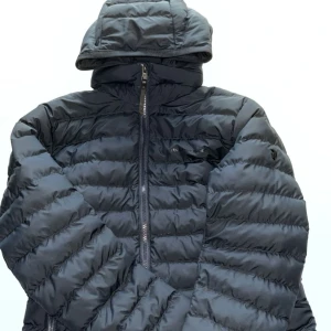 Svart dunjacka med huva från Peak Performance - Säljer en svart dunjacka från Peak Performance med huva och dragkedja framtill. Jackan har quiltad design och diskret logga på bröstet samt ärmen. Perfekt för kyliga dagar och enkel att matcha med olika outfits.
