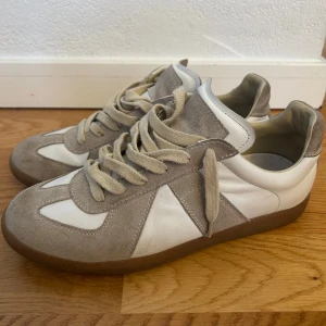 Maison Margiela Gats sneakers i mocka och skinn - Säljer ett par Maison Margiela Gats sneakers i beige mocka och vitt skinn med klassisk gummisula. Skorna har snörning och en stilren, retroinspirerad design som passar perfekt till streetwear och casual outfits.