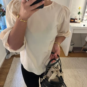 Beige blus med puffärmar och rosett i ryggen - Säljer en beige blus med puffiga ärmar och fina rosettdetaljer i ryggen. Blusen har rund halsringning och är tillverkad i ett mjukt material. Perfekt för dig som gillar romantiska detaljer och en stilren look.