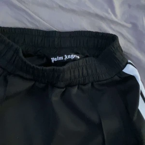 Svarta träningsbyxor från Palm Angels med vita ränder - Svarta träningsbyxor från Palm Angels med vita sidoränder och elastisk midja. Byxorna har logotypen broderad på ena benet och en sportig, rak passform. Perfekta för en streetwear-look.