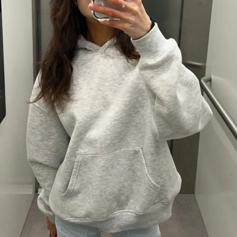 Säljer en ljusgrå hoodie med stor känguruficka och huva. På ryggen finns blå tryckt text . Oversize i modellen. Neuletakit & Villapaidat.