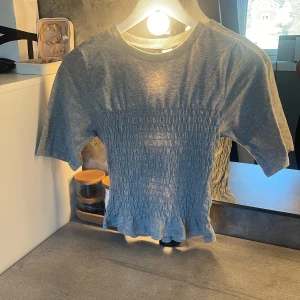 Grå smockad topp från H&M - Grå kortärmad topp från H&M med smockad midja som ger en snygg passform. T-shirten har rund halsringning och är tillverkad i mjukt bomullsmaterial. Perfekt för en avslappnad och trendig look.