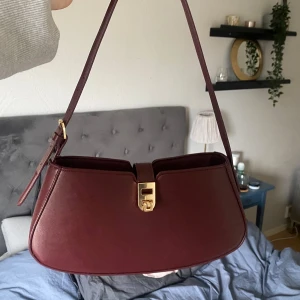 Vinröd axelväska med gulddetalj - Säljer en stilren vinröd baguette väska med justerbart axelband och snyggt guldfärgat spänne framtill🥰