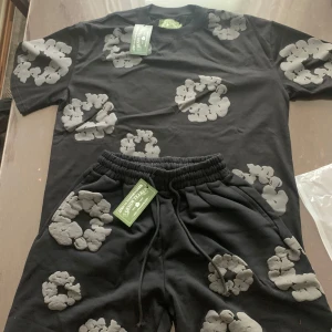 Svarta shorts med grått mönster från Denim Tears - Svarta shorts från Denim Tears med unikt grått, molnliknande mönster över hela plagget. Shortsen har resår i midjan och snörning för justerbar passform. Perfekta för en avslappnad och trendig stil.