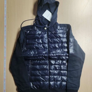 Mörkblå hybridjacka med huva från Moncler - Säljer en mörkblå hybridjacka från Moncler med glansig quiltad front och ärmar i mjuk bomull. Jackan har huva, dragkedja och en ficka med Moncler-logga på ärmen. Perfekt för lager-på-lager och snygg streetstil.
