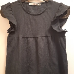 Mörkblå top med volangärmar. Strlk 140 - Mörkblå top med rund halsringning och fina volangärmar. Toppen har en lätt rynkad söm under bysten och är tillverkad i ett strukturerat tyg som ger en snygg look.