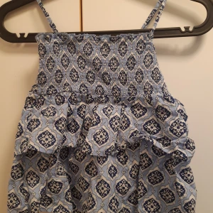 Blå mönstrad top med volanger från Gina Tricot - Säljer en blå klänning från Gina Tricot med smala axelband och volangdetaljer. Klänningen har ett vitt och mörkblått mönster och smock upptill för en bekväm passform. Perfekt för varma dagar!