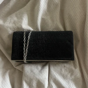 Svart glittrig clutch - Snygg svart kuvertväska med glittrigt tyg och silverfärgad kedja. Perfekt att bära över axeln eller i handen då man bara kan lägga in kedjan i väskan. Perfekt till bal eller student. Aldrig använd.