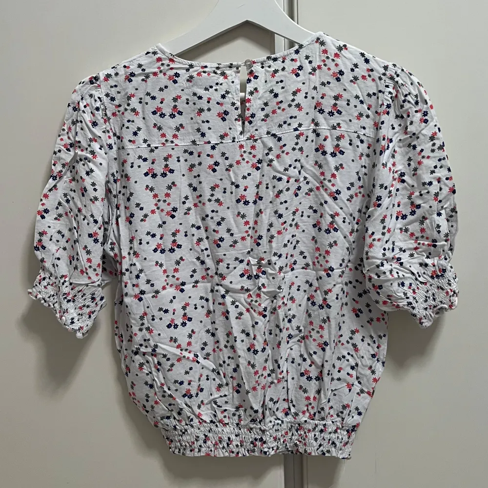 Söt vit blus med små blommor i rosa, blått och grått. Blusen har korta puffärmar med resår och resår i midjan för en feminin siluett. Perfekt för vår och sommar, lätt och luftig i bomull.. Yläosat.