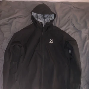 Svart skaljacka från Haglöfs - Säljer en svart skaljacka från Haglöfs med huva och dragkedja framtill. Jackan har minimalistisk design och är perfekt för utomhusaktiviteter. Lätt och smidig med diskret logga på bröstet.