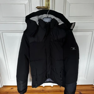 Jacka  - Säljer en svart pufferjacka från Jack and Jones  med huva och dragkedja framtill. Jackan har två fickor med dragkedja och en hög krage som skyddar mot vind. Perfekt för kyliga dagar.