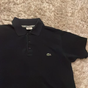 Mörk Blå pikétröja från Lacoste - Klassisk mörkblå  pikétröja från Lacoste med broderad logga på bröstet. Tröjan har korta ärmar, krage och knäppning med tre knappar framtill. Storlek L, kontakta vid funderingar.