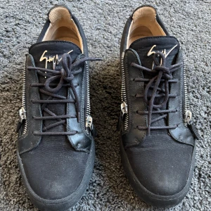 Svarta sneakers från Giuseppe Zanotti - Säljer ett par svarta sneakers från Giuseppe Zanotti med signaturlogga på plösen och coola silverfärgade dragkedjor på sidorna. Skorna har snörning och rund tå, samt en platt sula. Perfekta för dig som gillar streetstyle och exklusiva detaljer. Knappt använda.