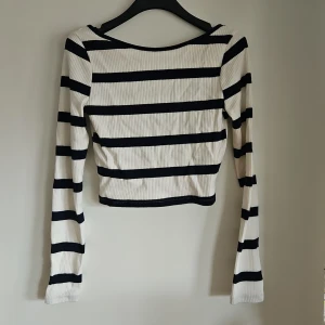 Randig croppad långärmad topp - Säljer en croppad långärmad topp med breda svarta och vita ränder. Toppen är ribbad och har en snygg öppen rygg som ger en cool twist. Perfekt för dig som gillar en trendig och avslappnad stil. Endast använd 1 gång.