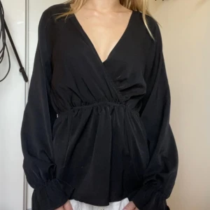 Svart omlottblus med v-ringning - Säljer en svart omlottblus med djup v-ringning och långa puffiga ärmar. Blusen har resår i midjan som ger en snygg siluett och vida ärmslut. Perfekt för dig som gillar en stilren och trendig look.