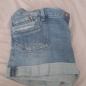 Blå jeansshorts med uppvikta ben - Snygga blå jeansshorts med klassisk femficksdesign och uppvikta ben. Shortsen har bälteshällor, kopparfärgade knappar och en bakficka med lock och knapp. Perfekta för varma dagar och en chill stil.
