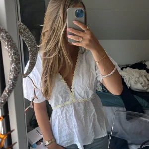 Vit blus med broderade blommor - Supersöt vit blus från Design by Si med v-ringning och broderade små blommor i gult och vitt längs halsringningen och midjan. Blusen har puffiga korta ärmar och är tillverkad i ett lätt, prickigt bomullstyg. Supersöt! Klicka görna på ”köp nu”🌼