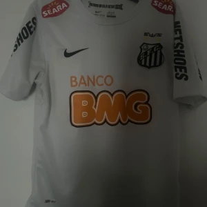 Santos Neymar tröja - Vit Santos fotbollströja med Neymar Jr och nummer 11 på ryggen. Tröjan har korta ärmar, flera sponsorloggor och klubbmärke på bröstet. Tillverkad i Dri-Fit material som andas, perfekt för match eller träning. Klassisk passform och snygg design.