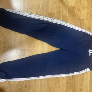 Marinblå mjukisbyxor från Gant - Snygga marinblå mjukisbyxor från Gant med vita sidor där det står GANT i blått. Byxorna har dragsko i midjan, ribbade muddar vid bensluten och är riktigt sköna för chill eller träning. Klassisk och sportig vibe.
