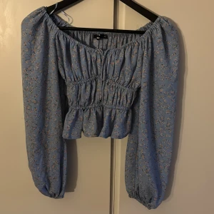 Blommig blå blus från BikBok XS - Supersöt blommig blus från BikBok i ljusblått med små vita och rosa blommor. Modellen är croppad med smockad midja, volangkant och långa puffärmar med resår. 