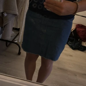 Mörkblå jeanskjol med rå kant - Säljer en mörkblå jeanskjol med rå, fransig kant nertill. Kjolen har klassisk femficksdesign, bälteshällor och dragkedja med knapp. Vet inte var de är ifrån eller storlek men skulle gissa på S