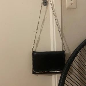 Svart axelväska med kedja - Snygg svart axelväska i skinn med silverfärgad kedja som axelrem och detaljer längs kanterna. Stilren och enkel design som passar till många outfits. Perfekt storlek för det viktigaste du behöver ha med dig.