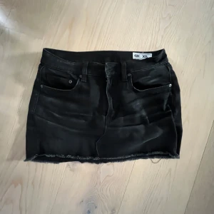 Svart låg midjad jeanskjol  - Svart jeanskjol  i storlek XS. Klassisk femficksmodell med bälteshällor och rå, fransig kant nedtill för en edgy vibe. Perfekt till sneakers eller boots och funkar året runt. Ljuset är dåligt, den är inte så vit.