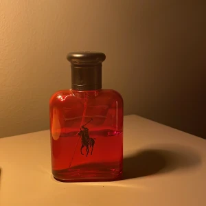 parfymflaska från Ralph Lauren söker byten - Snygg parfymflaska från Ralph Lauren i klarrött glas med svart logga av en polospelare. Flaskan har en stilren, rundad form och ett mörkgrått lock. Perfekt för dig som gillar klassisk och sportig design.