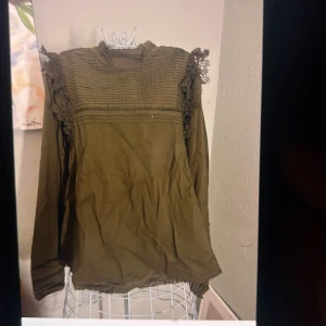 Olivgrön blus med spetsdetaljer - Snygg olivgrön blus med lång ärm och högt krage. Blusen har dekorativa spetsdetaljer på axlar och nederkant samt veckade partier upptill. Bra sick
