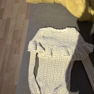 Vit stickad tröja med volang - Supersöt vit stickad tröja med lång ärm och volangdetalj över bröstet och axlarna. Tröjan har ett fint hålmönster och rund halsringning. Perfekt för dig som gillar romantisk och feminin stil.