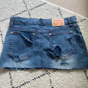 Ripped jeanskjol från Playh & Phil - Trendigt sliten jeanskjol från Playh & Phil med råa kanter och flera revor. Klassisk blå denim med rosa kontrastsömmar, två bakfickor och dragkedja framtill. Perfekt för dig som gillar en edgy och avslappnad stil.