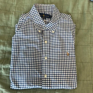 Blårutig skjorta från Ralph Lauren - Snygg blårutig skjorta från Ralph Lauren i slim fit-modell. Skjortan har klassisk krage, långa ärmar och knäppning framtill. Tillverkad i bomull och prydd med den ikoniska broderade loggan på bröstet. Perfekt för dig som gillar stilrena och tidlösa plagg.